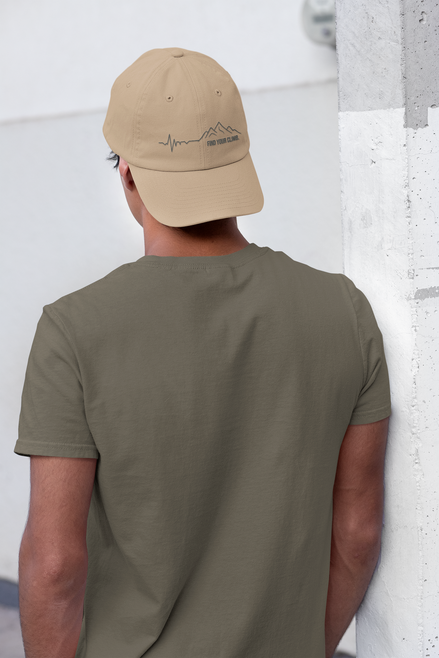 A man viewed from behind wearing an olive green t-shirt and a tan baseball cap with a black mountain logo on the back, leaning against a white wall.