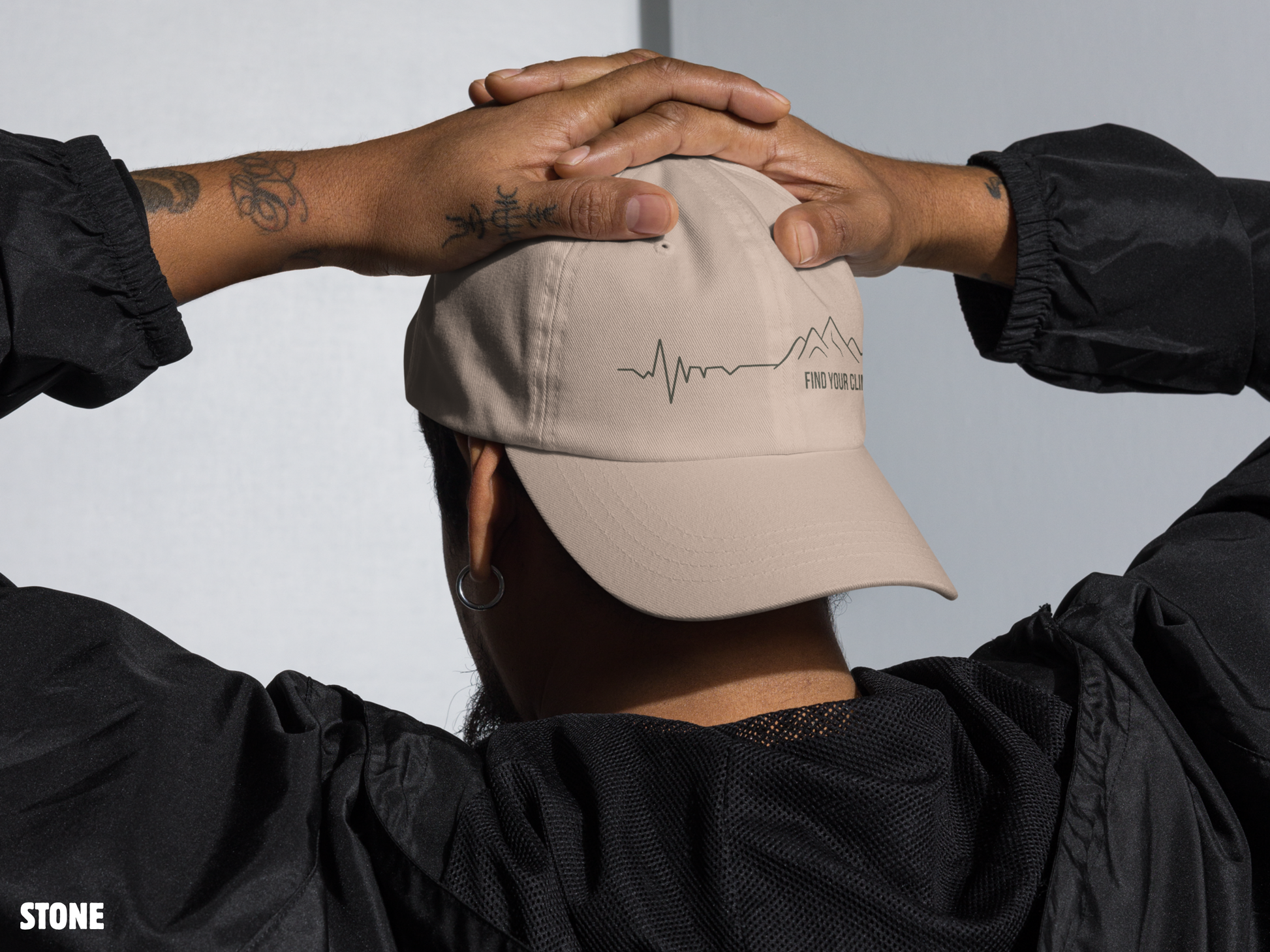 A person viewed from behind wearing a beige baseball cap with a black line-art mountain and heartbeat logo on the back, along with a black jacket. The person's hands are clasped behind their head against a light gray background.