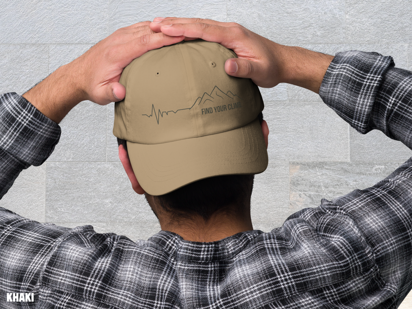 A man viewed from behind wearing a khaki baseball cap with an embroidered mountain and heartbeat line design on the front. His hands are on top of the cap, and he is wearing a black and white plaid flannel shirt against a light gray textured wall.